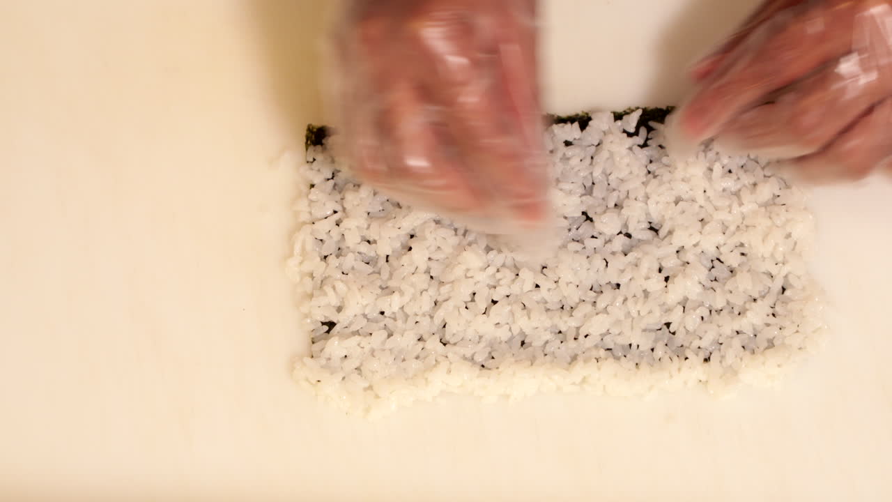 manos con guantes de plástico desechables preparando arroz de sushi en nori para rodar - toma cenital, cámara lenta