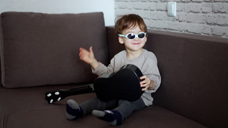 cute funny little kid wearing sunglasses sits on the sofa. adorable toddler holds a small guitar.