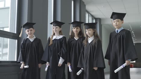 group of happy multiracial preschool students in mortarboard and gown. they are holding their diplomas and waving.