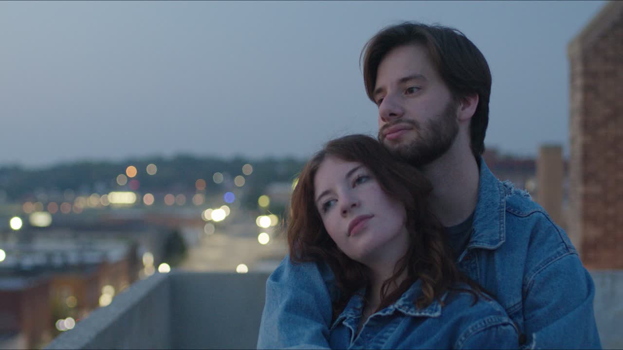 Couple wearing denim hold each other on a rooftop at night in an urban area