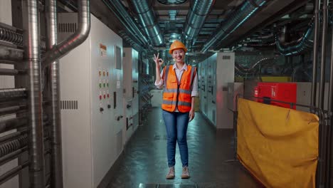 full body of asian female engineer with safety helmet smiling and showing okay gesture to the camera while standing in engine control room, work of electrical generators
