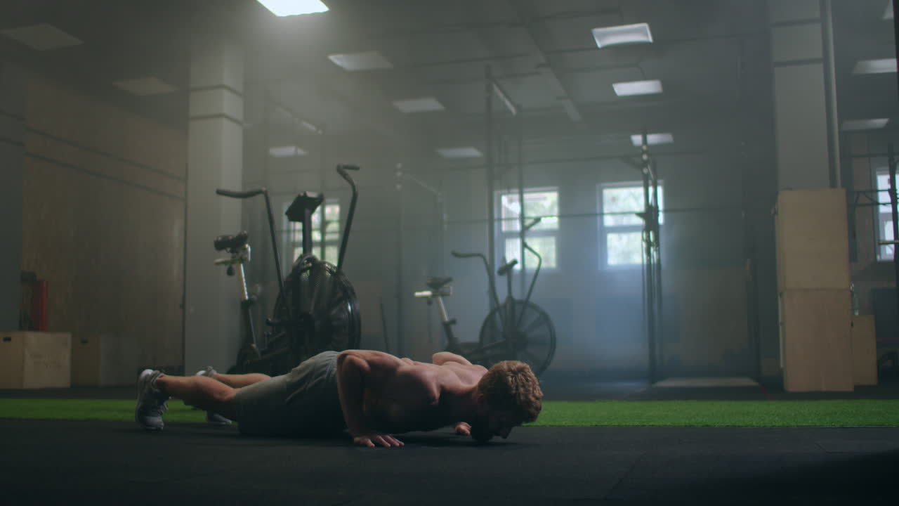 Uomo atletico, forte e in forma, in canotta sportiva e pantaloncini, sta facendo esercizi di piegamenti sulle braccia in una palestra industriale in stile loft con poster motivazionali. Fa parte del suo allenamento di cross fitness.
