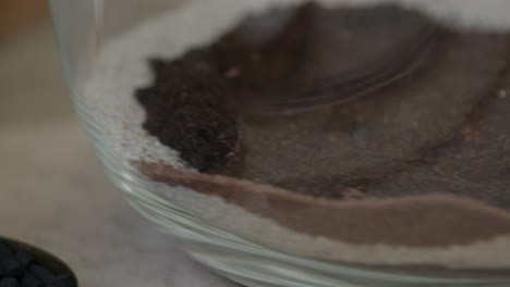 A-young-female-botanist-creates-a-tiny-live-forest-ecosystem-in-a-glass-terrarium---putting-brown-sand-layer---a-tight-close-up