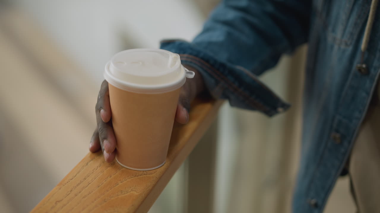 Close-up of hand holding paper coffee cup, resting on wooden railing in modern indoor setting with soft background, casual moment of calm, enjoyment, and relaxation