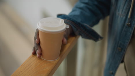 close-up of hand holding paper coffee cup, resting on wooden railing in modern indoor setting with soft background, casual moment of calm, enjoyment, and relaxation