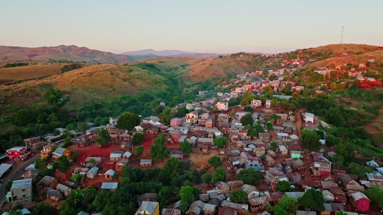 Drone footage of colourful hillside village surrounded by rolling hills and greenery in Madagascar, showing traditional architecture and red earth terrain at golden hour.