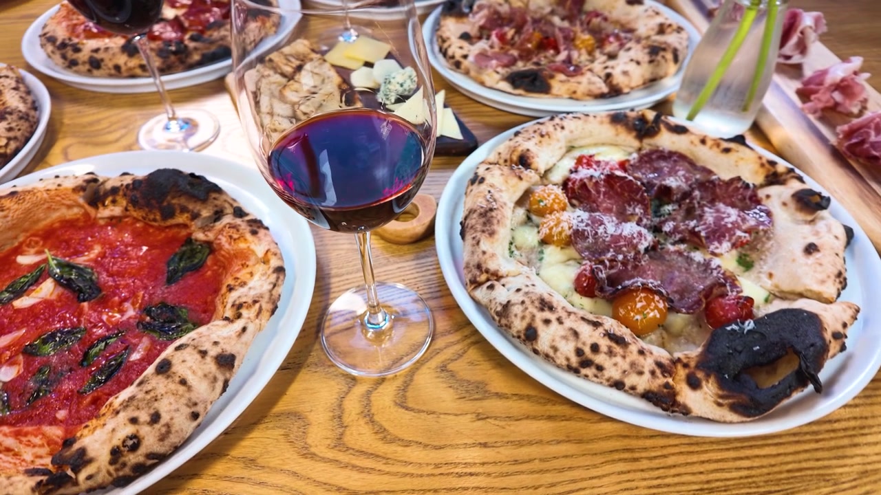 High-angle panning shot of gourmet pizzas, red wine, and appetizers on a wooden table.