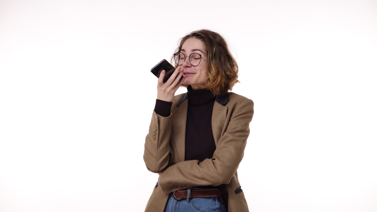 Beautiful brunette woman in stylish casual clothes and glasses records a voice message on her mobile phone on white background in studio. Talking emotionally to her friend and smiling