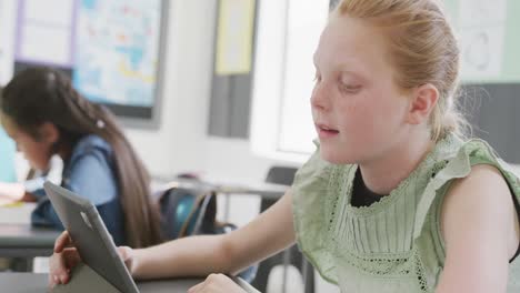 Video-of-frustrated-caucasian-schoolgirl-sitting-at-desk-using-tablet-in-diverse-class,-copy-space