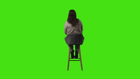 rear view full length shot of casually dressed young woman sitting on stool and waiting against green screen 1