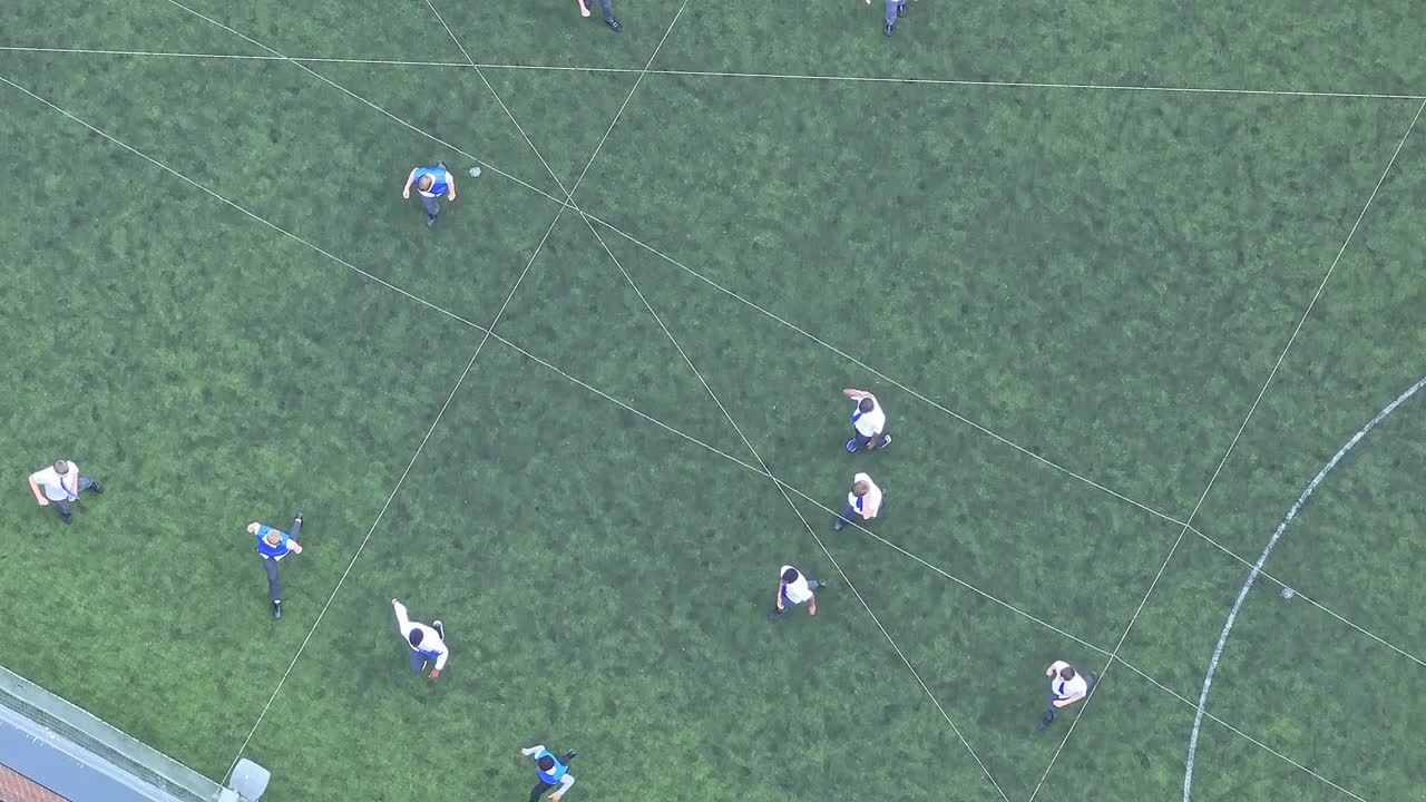 An aerial shot captures a soccer game in progress with players scattered across the green field marked by white lines. The central focus is on the ball and player positions, highlighting the dynamics