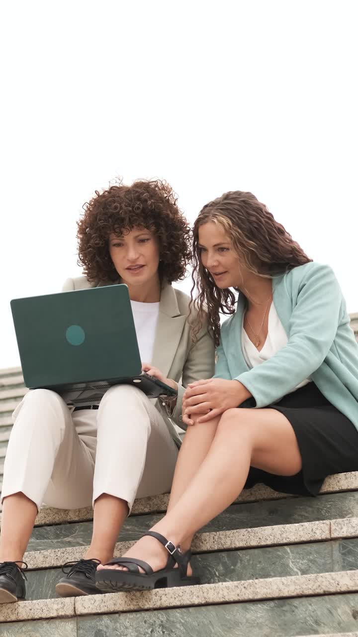 Businesswomen celebrating success using laptop on stairs. Vertical