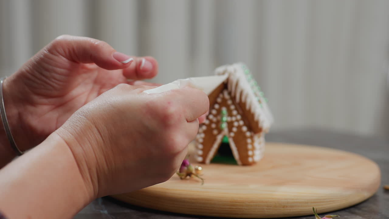 primer plano de una persona decorando una casa de pan de jengibre con glaseado, ligeramente borroso en el fondo en una bandeja de madera, manos aplicando cuidadosamente detalles intrincados a la estructura festiva de las galletas