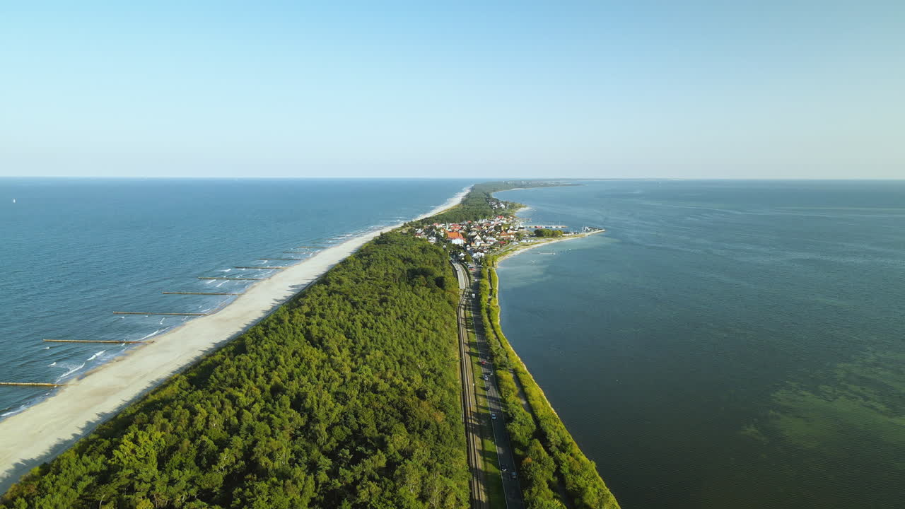 Luftoptagelse i kredsløb over europæisk havstrand på smal landtange med lille by og skov