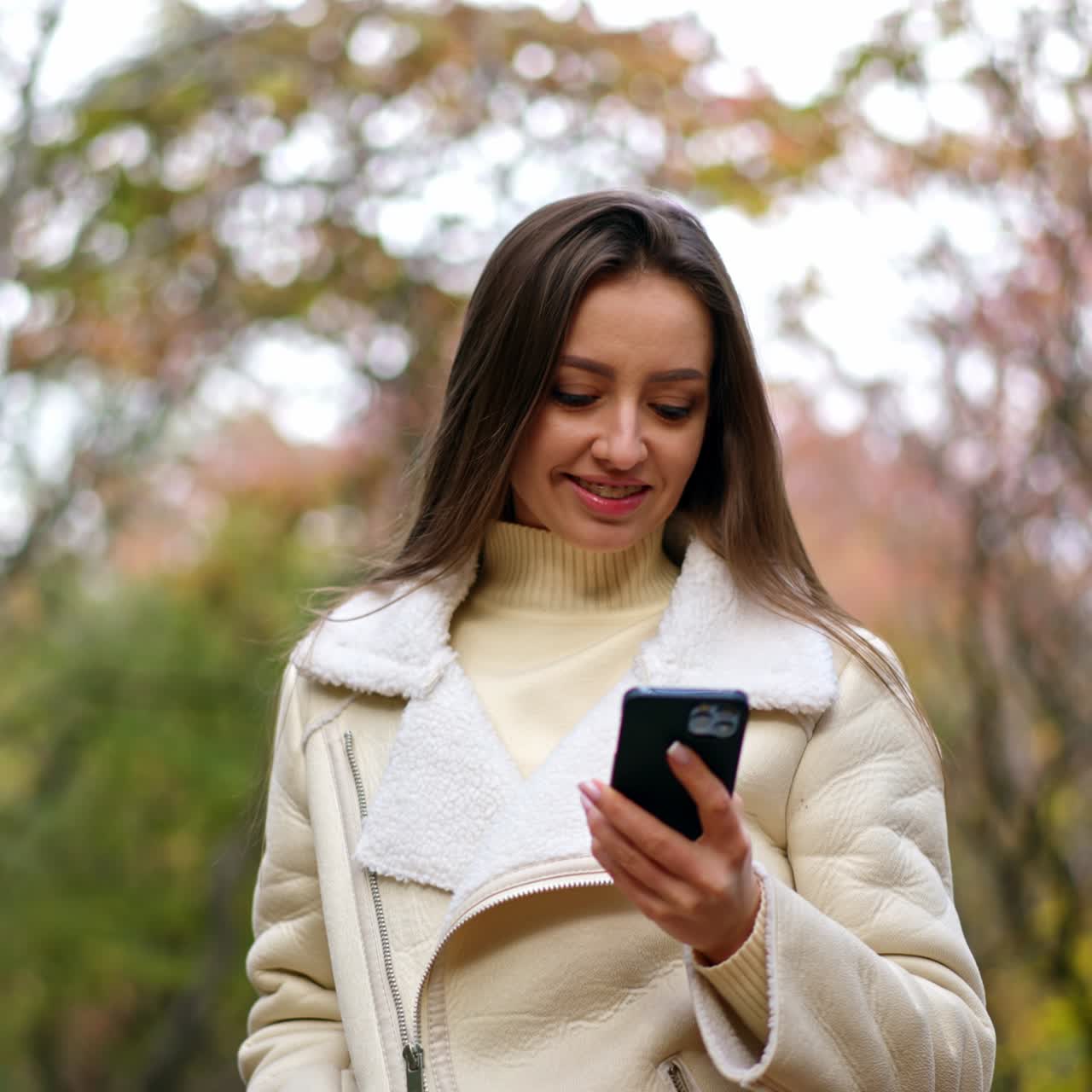 Beautiful smiling Caucasian lady speaks on the phone walking by the park. Lady goes looking at phone and then tries to take a picture of autumn trees