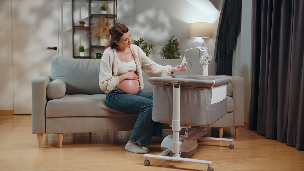 Pregnant woman sits on sofa touching belly near bedside bassinet with mobile pregnant woman with