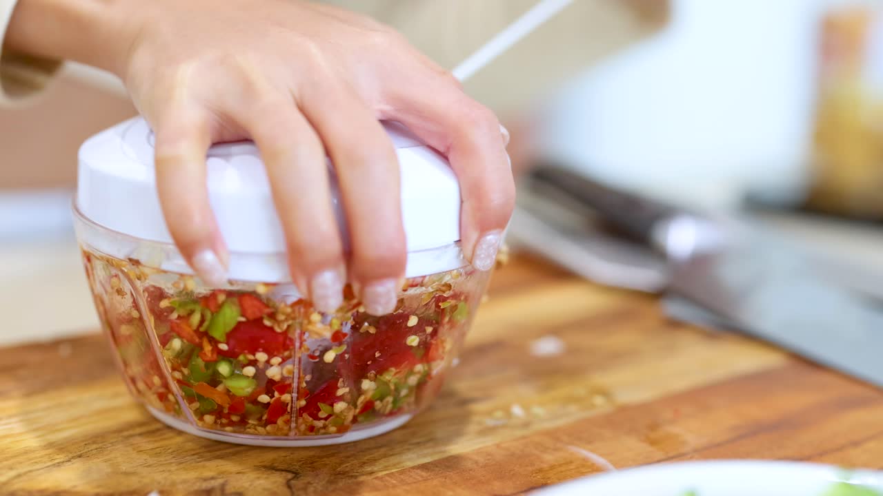A hand operates a manual food processor, chopping colorful vegetables on a wooden surface in bright lighting