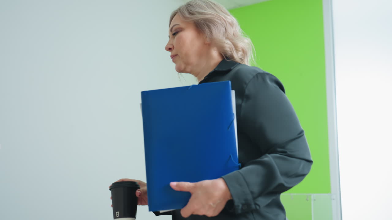Professional lady walks into office carrying document folder and tea cup, preparing to start work as she approaches desk in bright modern workspace with minimal decor and natural lighting