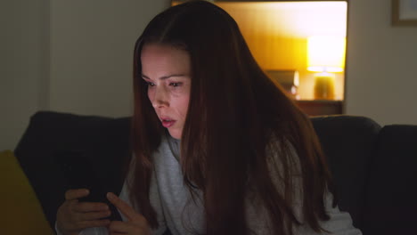 anxious woman sitting on sofa at home at night looking at mobile phone concerned about social media or bad news 2