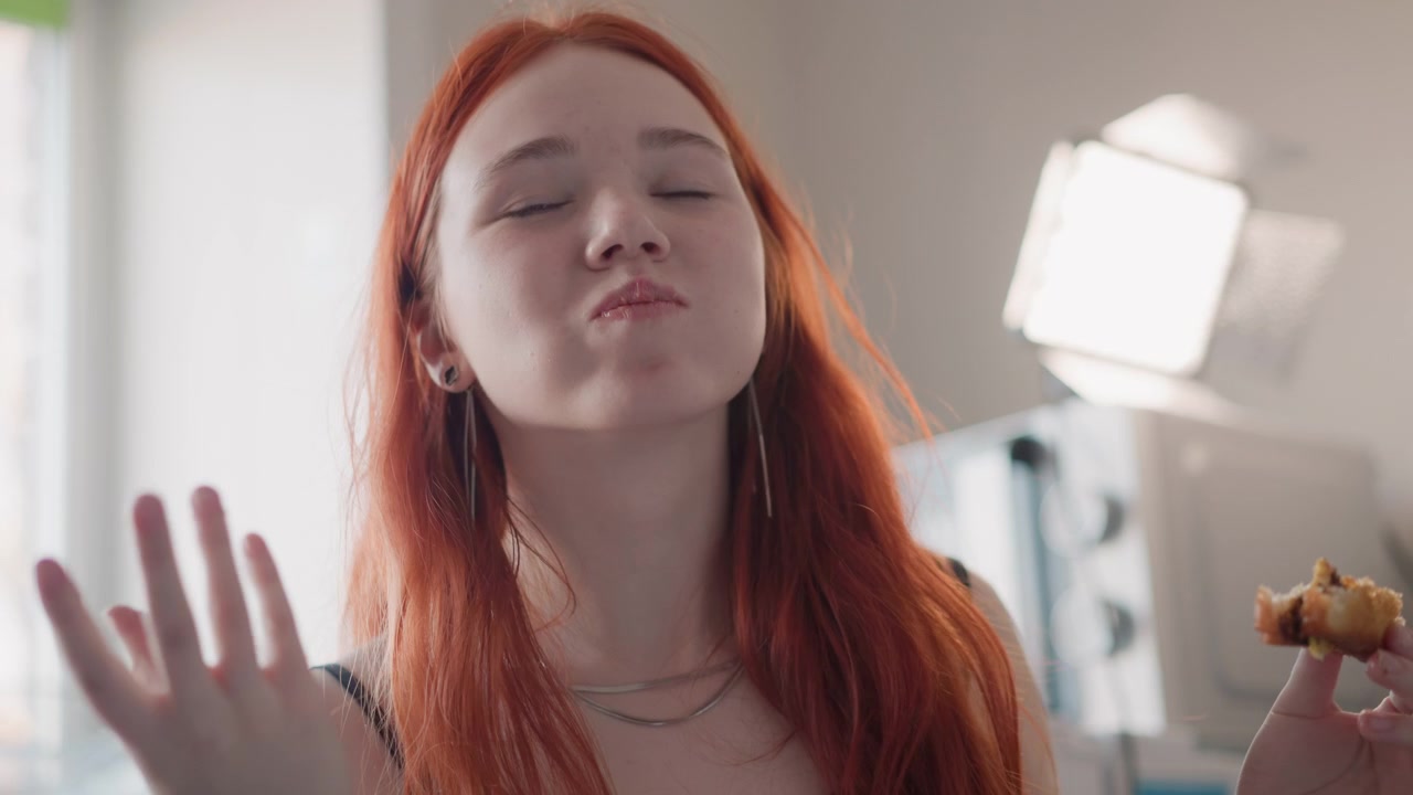 Lady relishes delight. Joyful woman with fiery hair savors dessert beneath gentle illumination. Woman with striking scarlet hair delightfully indulges in pastry under soft glowing light