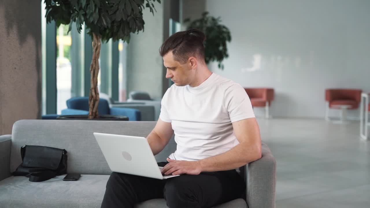 ⁇  티셔츠 를 입은 청년 이 비즈니스 센터 의 배경 에 앉아 노트북 뒤 에 피곤 하게 앉아서 일 하고 긴장감 있게 작업 을 끝내고 있는 장면