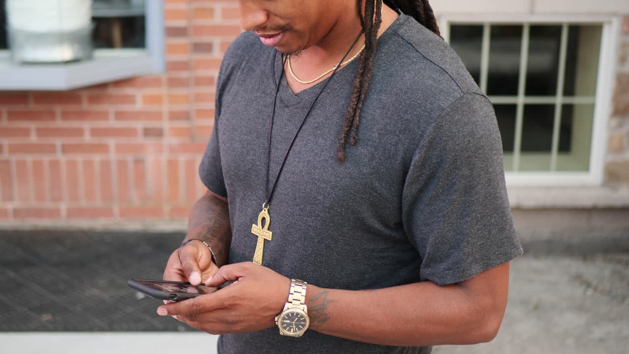 African American man with dreadlocks on his phone texting while wearing a gold watch and necklace, in a grey v neck t-shirt