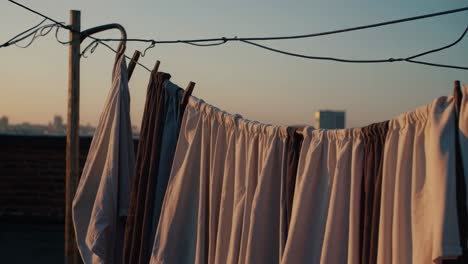 woman stands on a rooftop at sunset, observing clothes drying on a clothesline, with a city skyline in the background, creating a peaceful urban scene