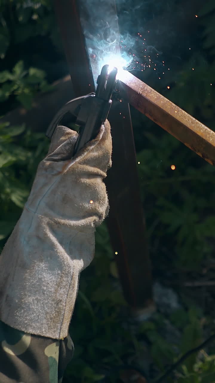 closeup handyman in protective work wear welds rusty carcass of country house fence in bushes