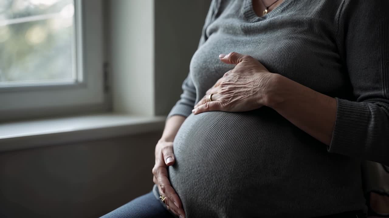 Donna incinta che si accarezza e si strofina la pancia, seduta vicino alla finestra, con un maglione grigio.