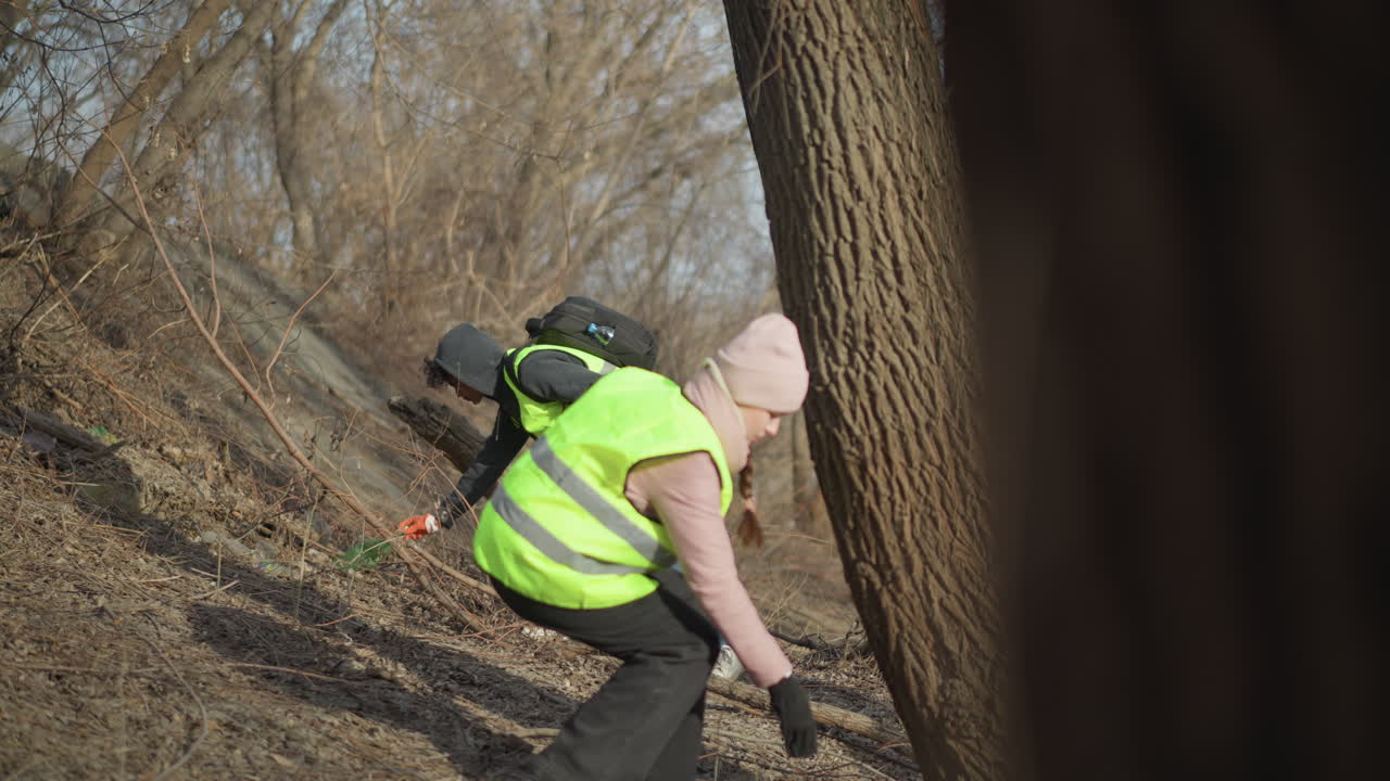 Two volunteers wearing reflective safety vests and gloves collecting trash in blue bags during outdoor cleanup along riverbank, removing litter to protect environment, reduce waste