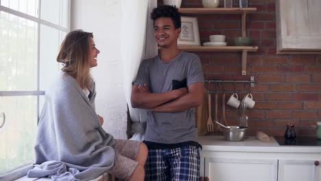 Attractive-girl-is-sitting-on-the-window-sill-covered-in-a-blanket-and-laughing-together-with-her-boyfriend.-Handsome