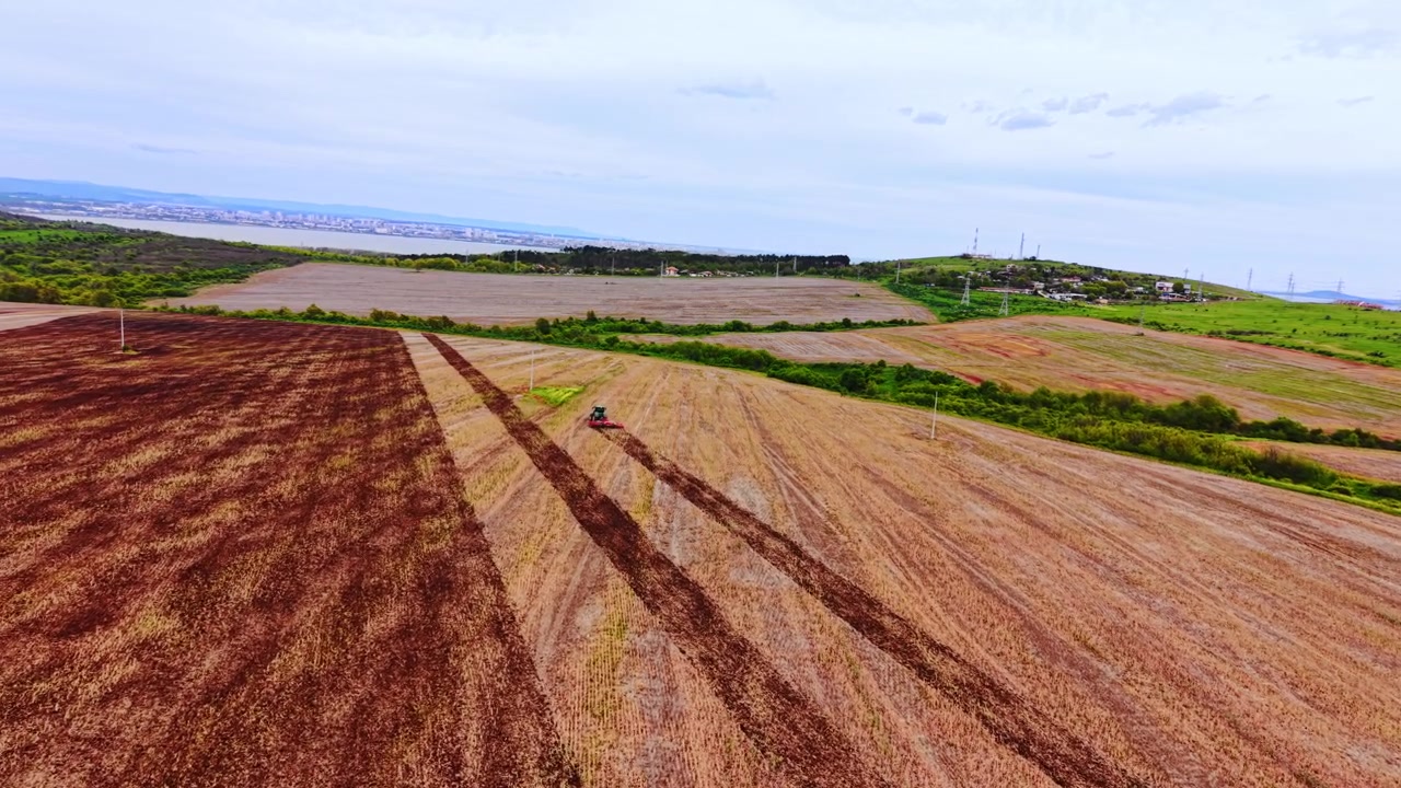 Pemandangan udara luas ladang yang telah dipanen. Sebuah traktor mengolah lahan pertanian cokelat kemerahan dengan teluk dan kota terlihat di baliknya