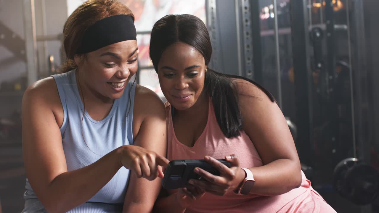 Diverse female friends flexing arm at gym, capturing and reviewing smartphone selfie and laughing