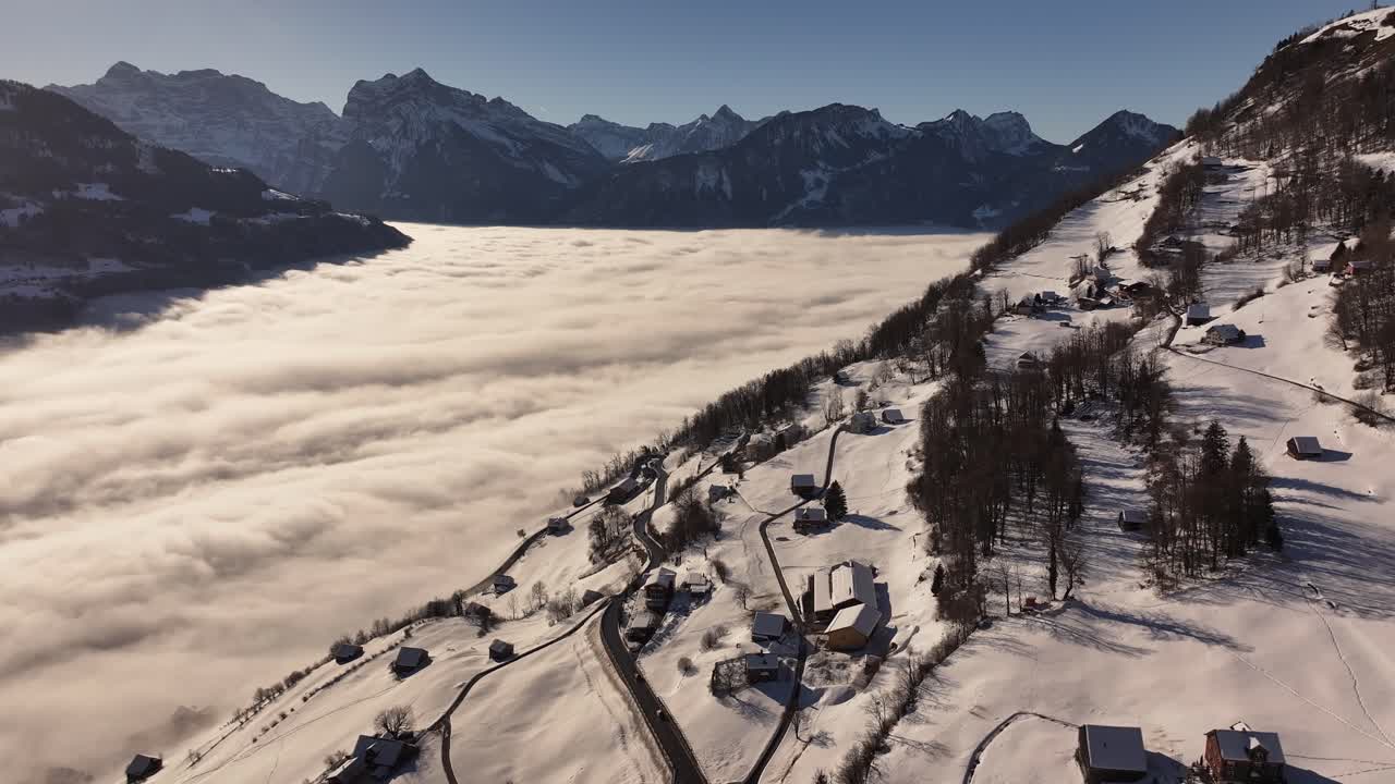 Aerial view of Amden, Switzerland, with a breathtaking sea of fog blanketing the valley, while snow-covered homes and rolling hills contrast against the distant alpine peaks.