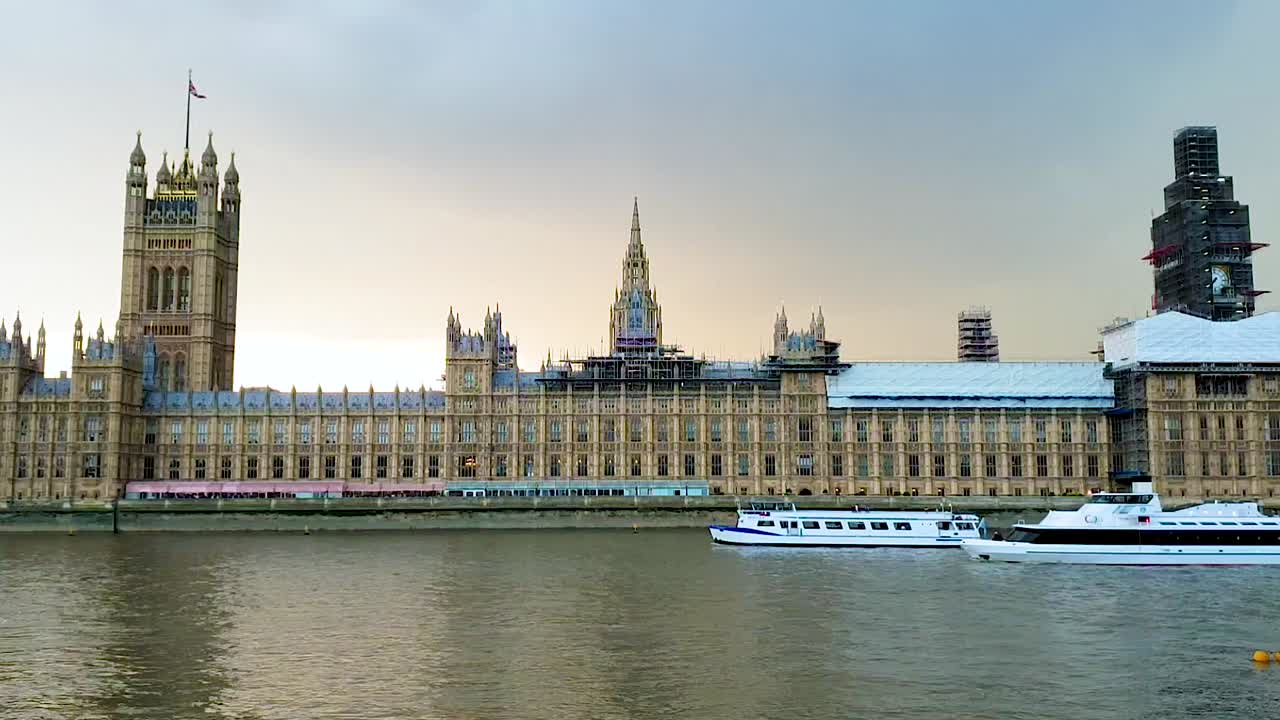 Slow motion footage showing boats moving along River Thames beside Houses of Parliament and scaffolded Elizabeth Tower in Westminster, London, under soft evening sky and calm water