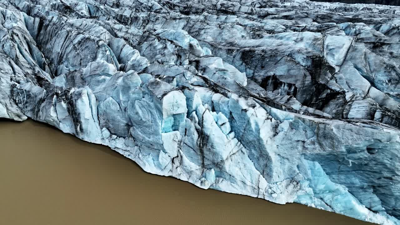 Drohnenansicht einer blau-weißen Gletscherwand, die auf einen braunen Schlicksee trifft, mit dunklen Ascheschichten, Gletscherspalten und scharfen Graten entlang der schmelzenden Küstenlinie