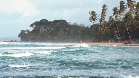 Tribal-Gathering-festival-at-Playa-Chiquita-Beach-in-Panama-with-people-walking-the-shore-on-windy-day,-Handheld-wide-shot