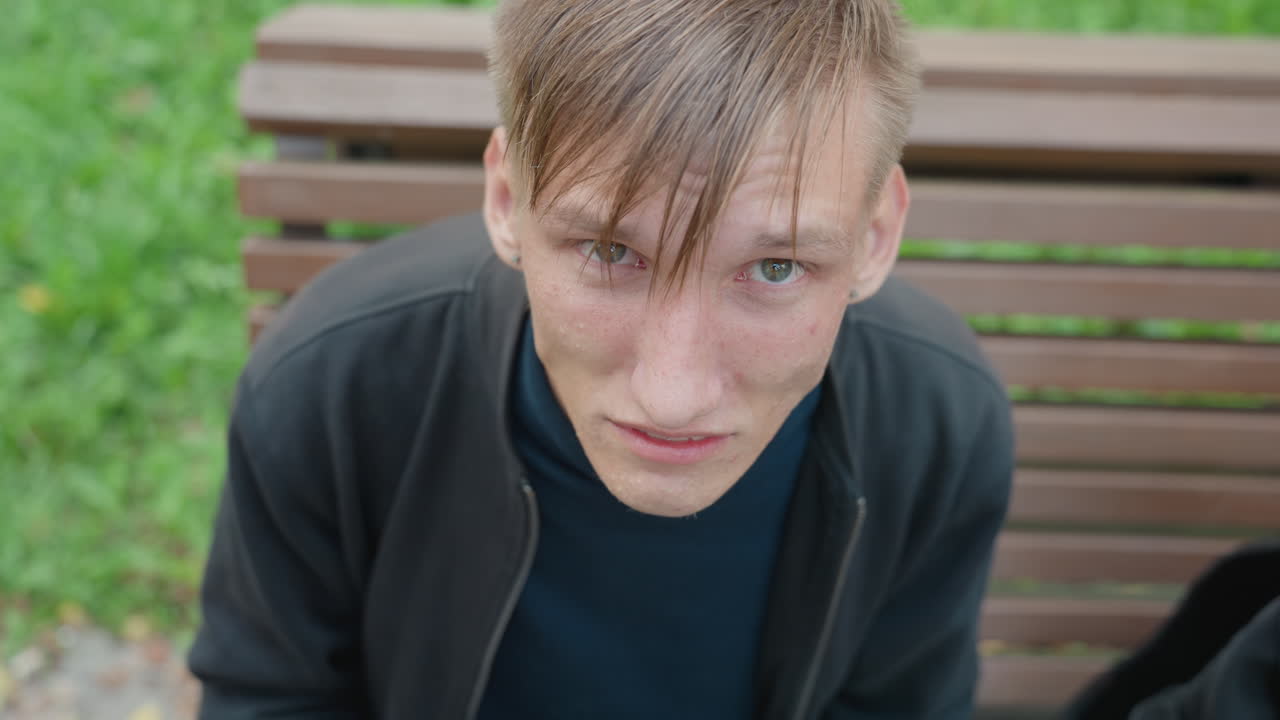 Homme fixant la caméra depuis un banc de parc, cheveux mouillés, regard intense, confrontation directe, environnement urbain brut, lumière du jour, gros plan, expression brute, sac à dos à côté de lui, arrière-plan herbeux, rôles de skateur, poète de rue, acteur