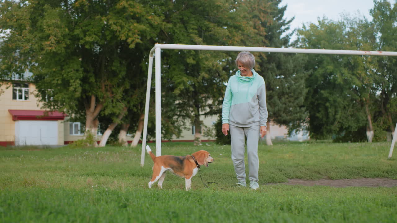 Trainer smiling while doing squats outdoors near goal post with beagle dog beside her on grassy field surrounded by trees showing fitness, active lifestyle, happiness, and healthy outdoor activity