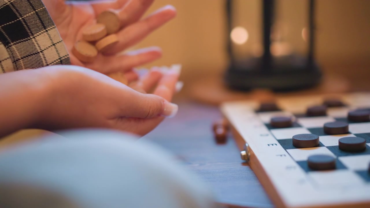 Primo piano di mani che tengono piccoli oggetti rotondi, con una scacchiera e un'illuminazione soffusa sullo sfondo. Una persona indossa un tessuto a quadri mentre afferra o mostra delicatamente gli oggetti in un ambiente interno