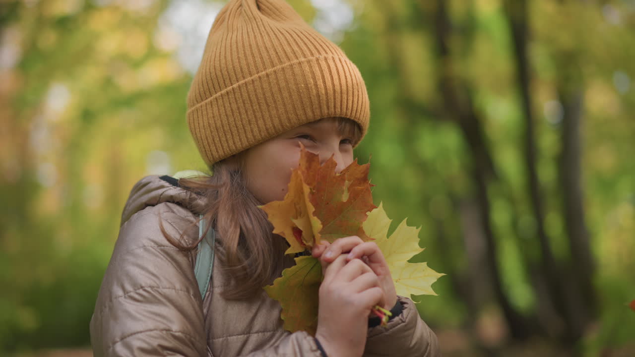 young girl wearing brown jacket and mustard beanie playfully holds autumn leaves close to face while smiling in golden forest, background