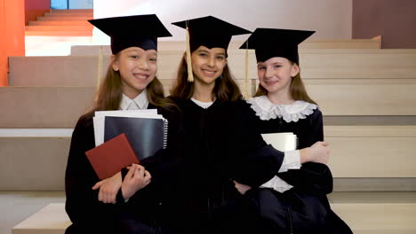 portrait of three happy little girls in cap and gown holding notebooks and looking at the camera while sitting on stairs at the preschool graduation ceremony