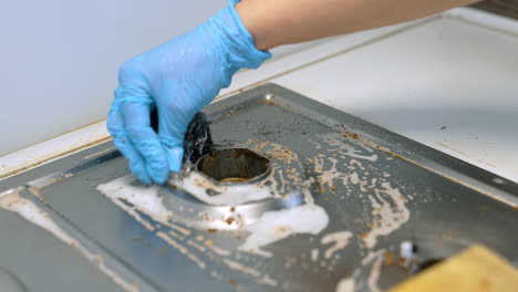 cleaning a kitchen sink with soap. a person in a blue glove scrubs a dirty kitchen sink drain to remove grime and food residue