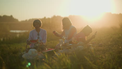 two sisters enjoying a summer picnic with fresh watermelon while one picks out seeds and the other takes a bite, beside them, a young girl is bent down, savoring her slice