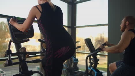 un grupo de personas en el área de cardio de la sala de gimnasia. el hombre usa la bicicleta de ejercicio elipsoidal de la mujer. toma de steadycam