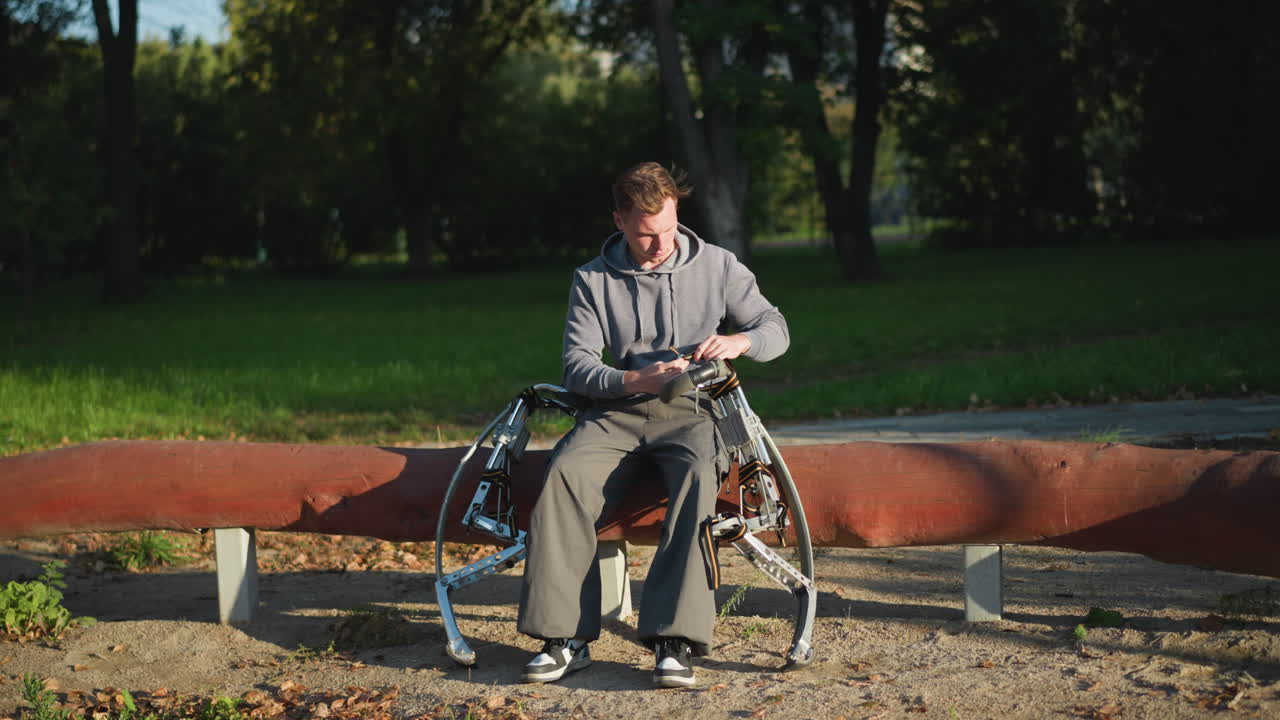 회색 후디를 입은 젊은이가 공원 벤치에 앉아서 은 날에 금속 봄 기둥을 조정하고, 푸른 풀과 나무로 둘러싸여, 평화로운 야외 환경에서 집중된 표현으로