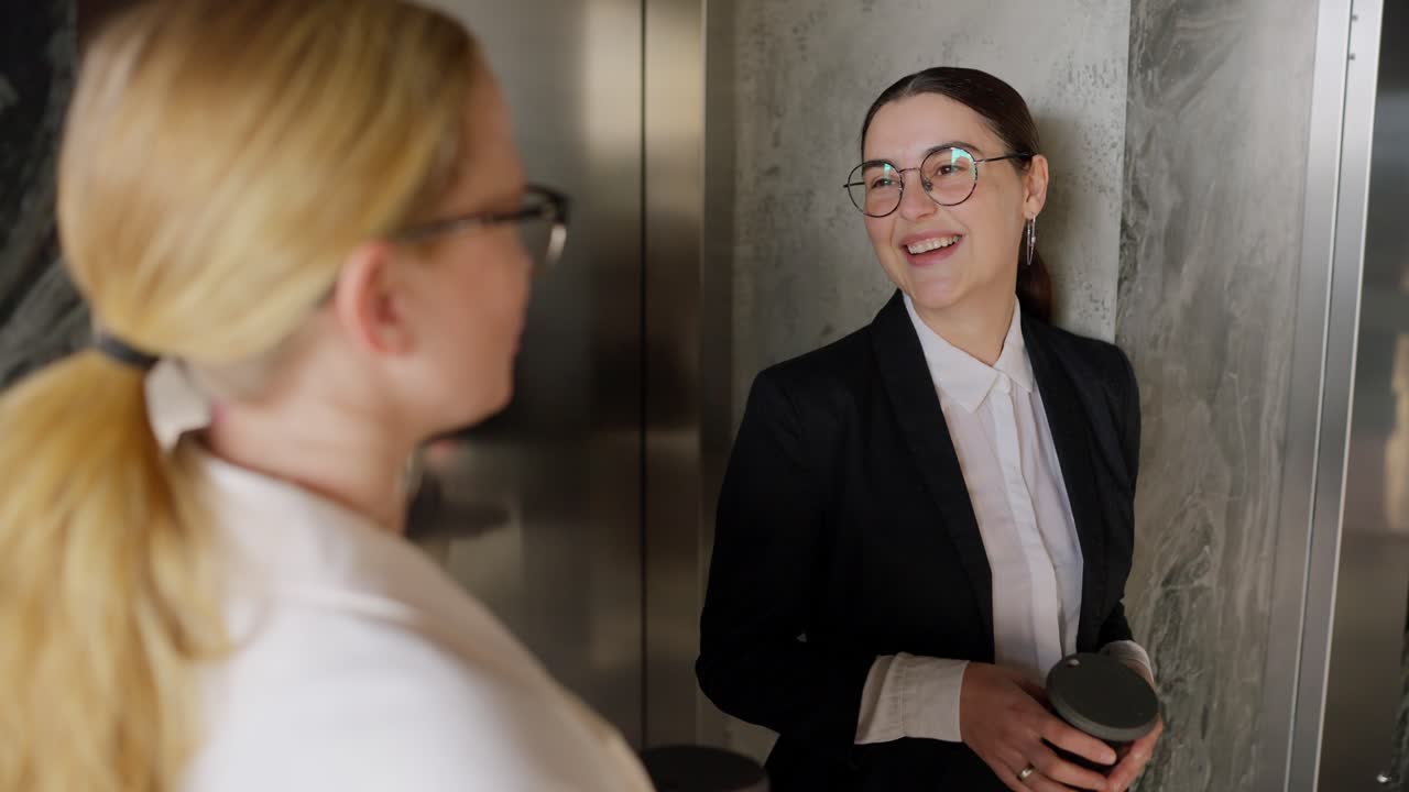 Over the shoulder A confident brunette girl in round glasses and a business suit communicates with her middle-aged blonde colleague during a short break and drinking coffee in a modern office