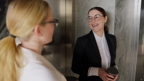 sobre el hombro una confiada chica morena con gafas redondas y un traje de negocios se comunica con su colega rubia de mediana edad durante un breve descanso y bebiendo café en una oficina moderna