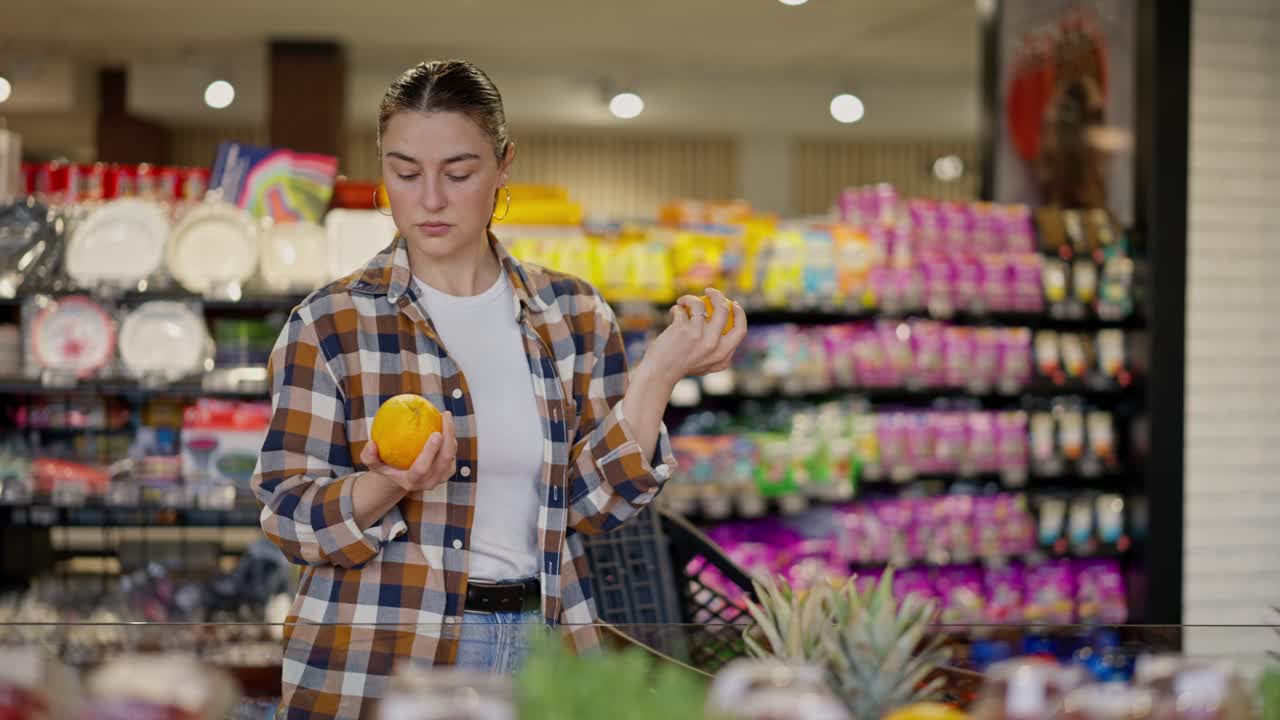 una chica morena con una camisa a cuadros elige una de las dos naranjas en el departamento del supermercado mientras compra