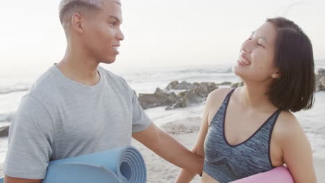 happy biracial couple holding yoga mats at beach, in slow motion